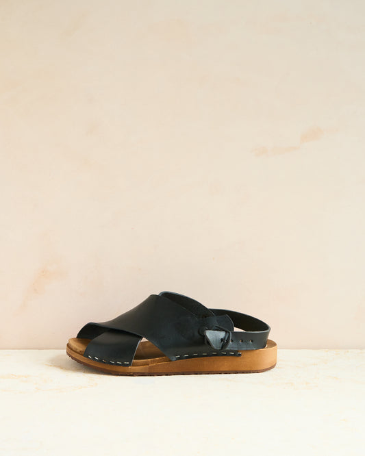 A black sandal with a flexible clog base and stapled leather uppers featuring an adjustable buckle strap at the back, set against a wooden background.
