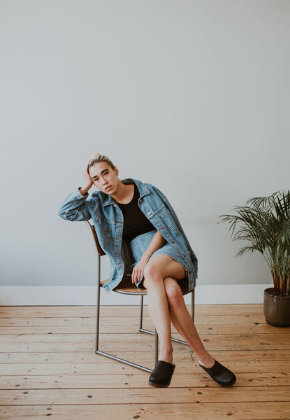 girl sits on a chair wearing a denim jacket and skirt with black low classic style swedish clog
