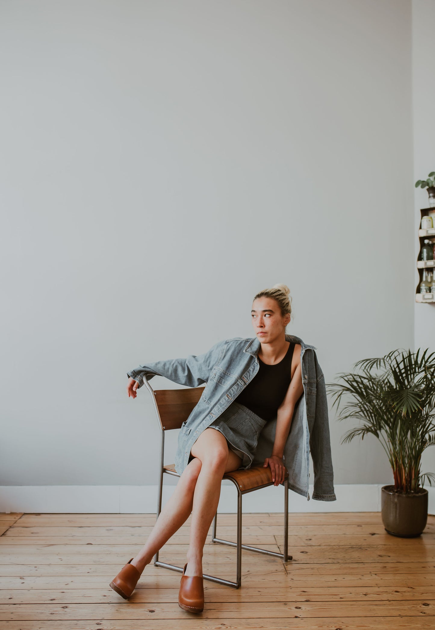 woman poses sitting on a chair wearing a denim jacker and skirt and tan cumin low heel classic style swedish clog with a covered back