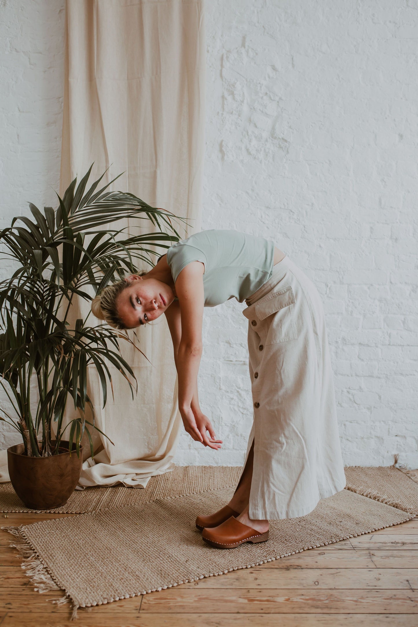 woman poses wearing a sage green tshirt, a raw beige coloured linen skirt and cumin orangey tan brown low classic style swedish clogs