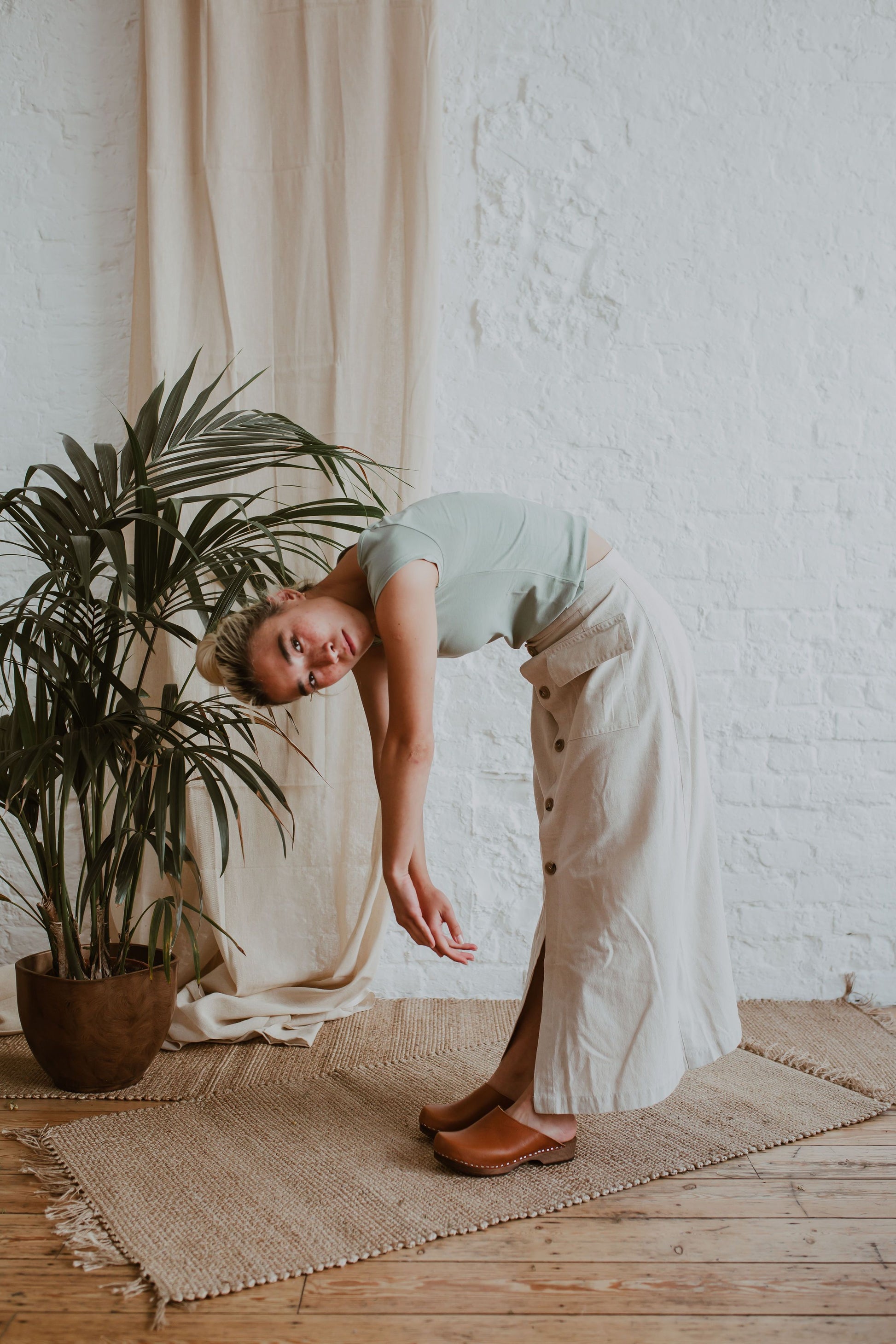 woman poses wearing a sage green tshirt, a raw beige coloured linen skirt and cumin orangey tan brown low classic style swedish clogs