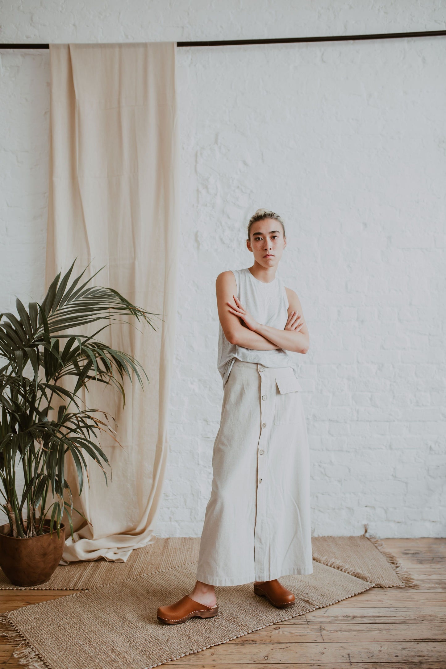 woman stands wearing a cream sleeveless top, a raw beige coloured linen skirt and cumin orangey tan brown low classic style swedish clogs