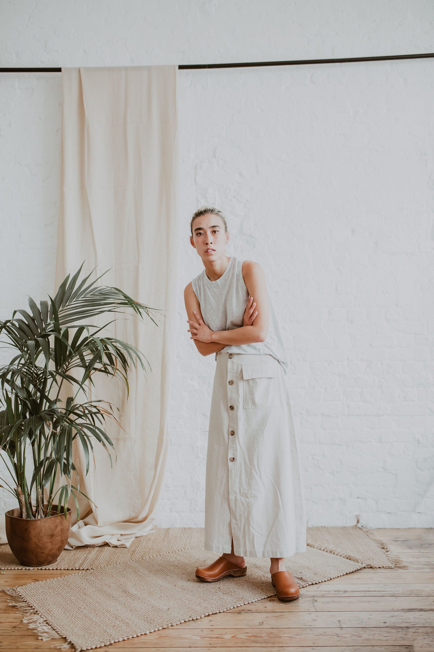 woman stands wearing a cream sleeveless top, a raw beige coloured linen skirt and cumin orangey tan brown low classic style swedish clogs
