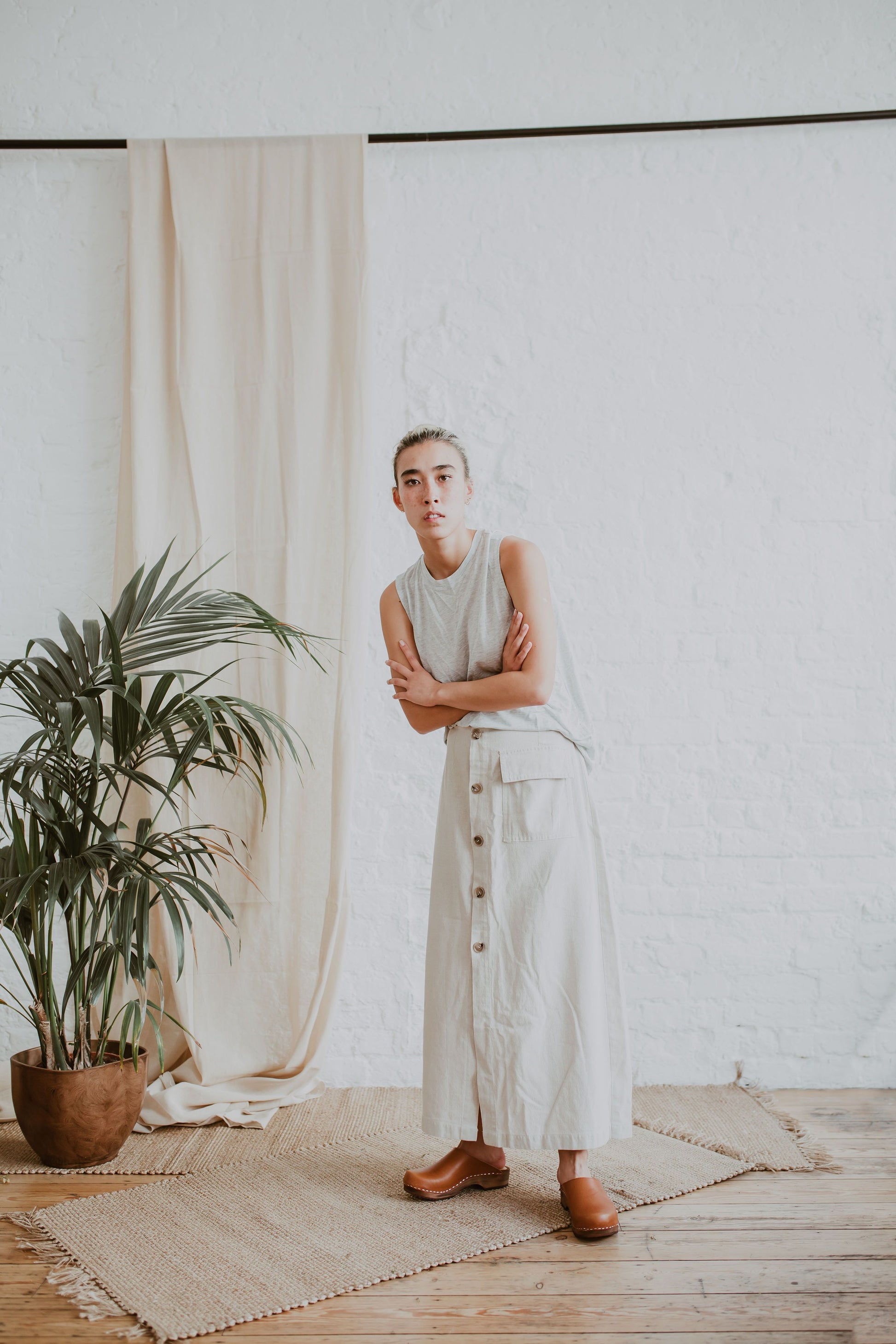 woman stands wearing a cream sleeveless top, a raw beige coloured linen skirt and cumin orangey tan brown low classic style swedish clogs