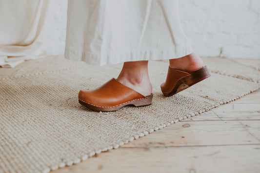 close up of woman's feet wearing a raw beige coloured linen skirt and cumin orangey tan brown low classic style swedish clogs